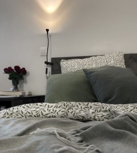 Bild på säng med grön filt och gröna örngott. Blommor, ljus och böcker på nattbordet bredvid