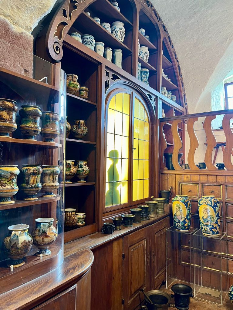Photo of a wooden shelf with many ceramic jars. The jars are mostly white but have blue drawings and inscriptions. In a cut out of the shelf a fake window can be seen with a silhouette of a lady.