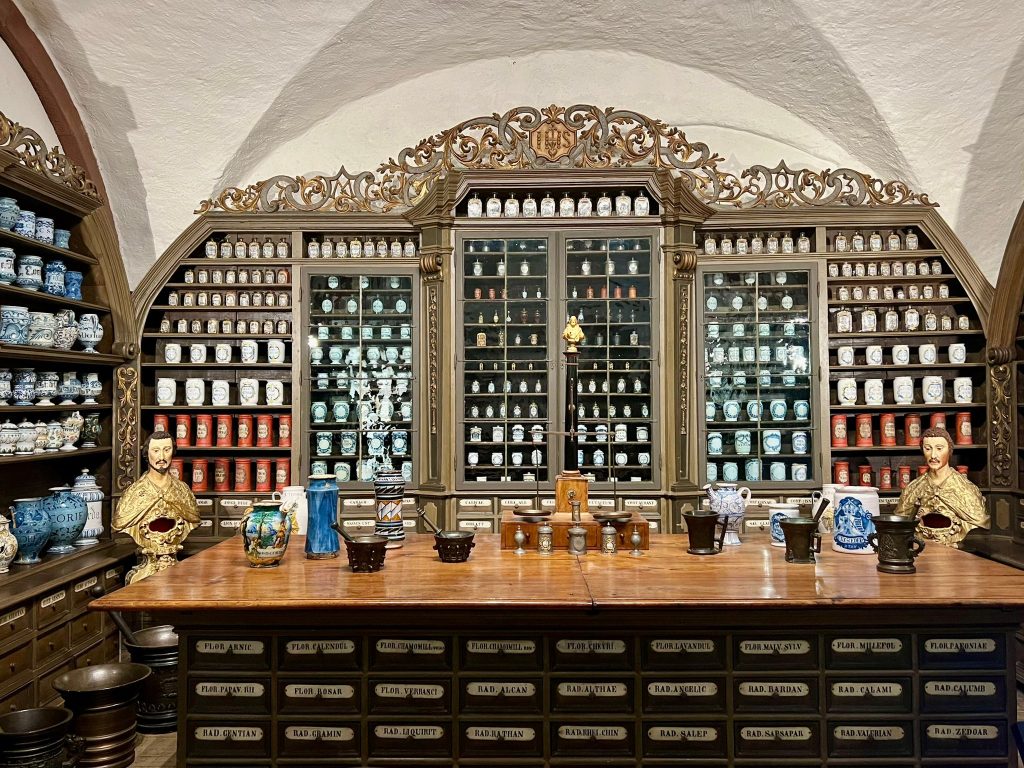 A room covered in wooden shelves. The shelves all have white and blue jars on them. In the middle of the room there is a waist height dresses with various ceramic objects on it.