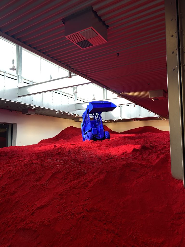 Konstverk i form av ett rum fyllt med röd jord och blå grävskopa på.