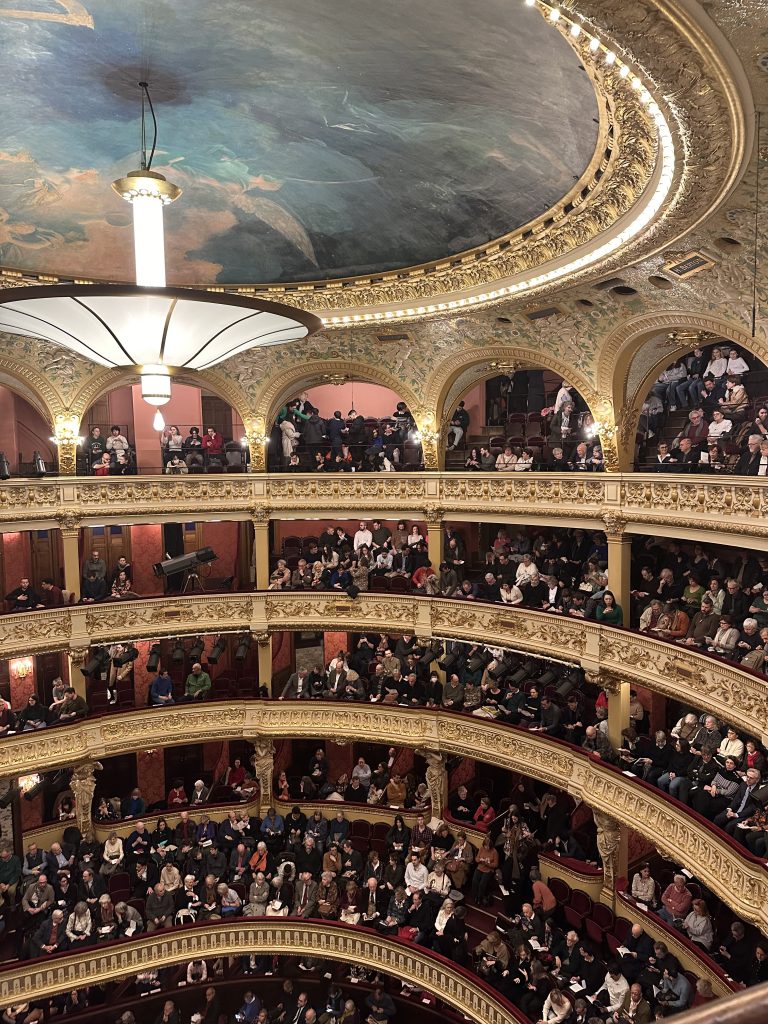En bild på en salong i ett operahus med gulddetaljer och färgglatt tak. Operahuset är fyllt med folk