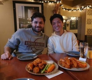 Two men looking at the camera in a bar with two plates of chicken wings in front of them on the table.