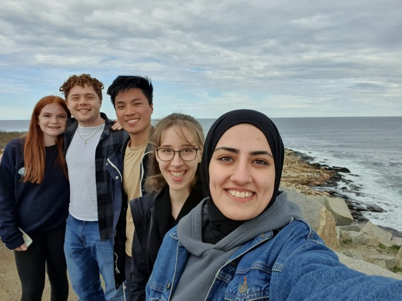 Group of people standing in a line smiling at the camera on the coast with the sea in the background.