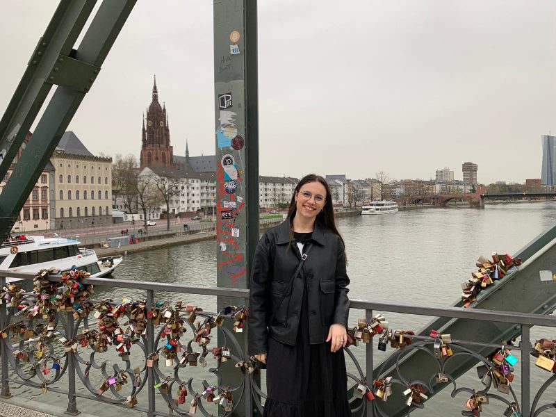 Photo of a girl smiling and dressed in black. She is on a bridge with locks hanged onto it. A church and the river are in the background.