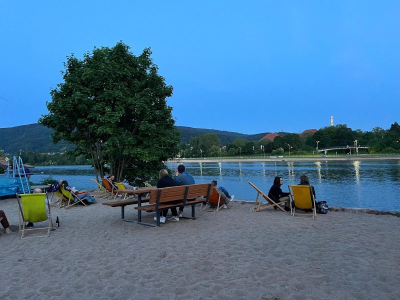 Photo of a sandy beach by a river. There is a green tree, people sitting on benches and chairs and the sky is a dark blue. It is evening.