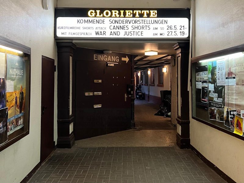 Photo of an entry to the cinema. At the top of the door there is a large lit up pannel with the text GLORIETTE and the names and schedule of the films currently being rolled. The door is a dark colour and has written EINGANG on it. On the walls of the hallway leading to the door there are movie posters.