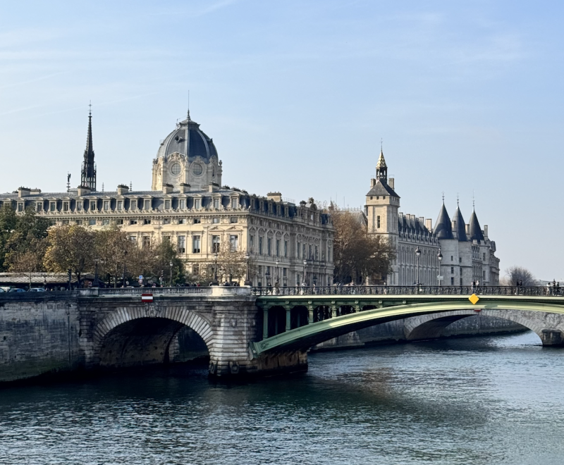 En vy över floden Seine i Paris med en grön bro i förgrunden och den historiska byggnaden Palais de Justice i bakgrunden, omgiven av träd och arkitektur från olika epoker