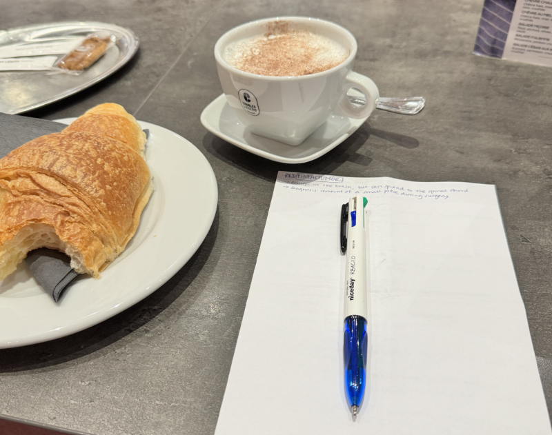 Croissant, cappucino och penna med anteckningsblock på ett grått bord.