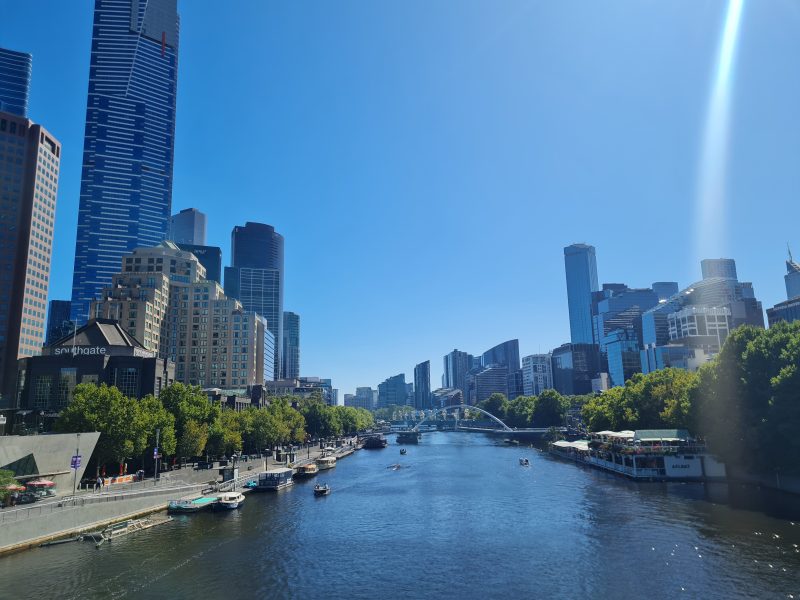 En panorama av Melbournes skyskrapor till höger och vänster med Yarra river i mitten och en vacker blå himmel.